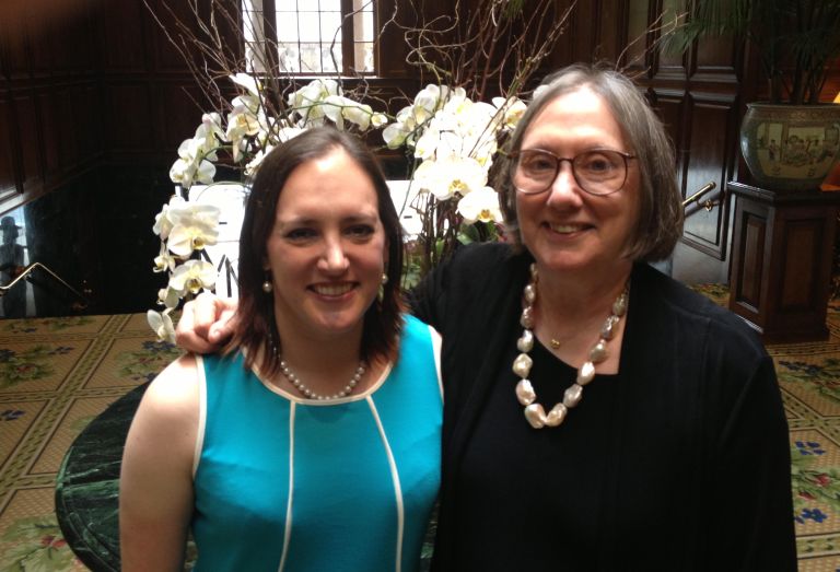 Two women in front of artificial orchids in a dark room. Left is Sarah Cook, a woman with shoulder-long brown hair. She wears a blue dress and pearl
earrings and necklace. Next to her is Dr. Delaney, a woman with grey hair and glasses wearing a black shirt.