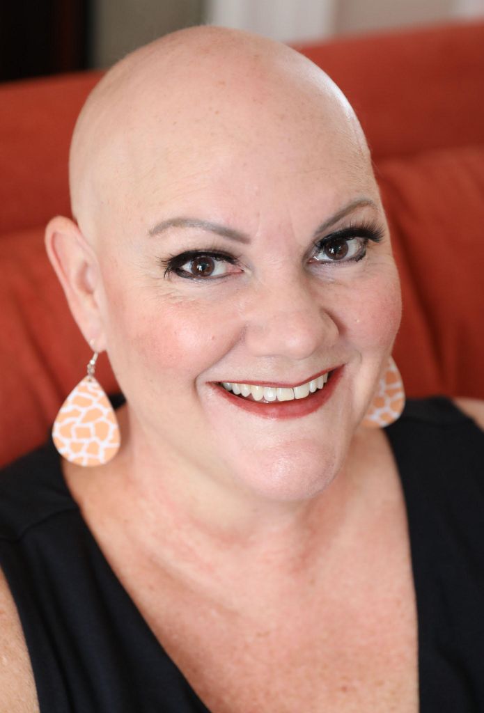 A woman with a bald head and large orange earrings. She sits on a couch and wears red lipstick and a black shirt. 
