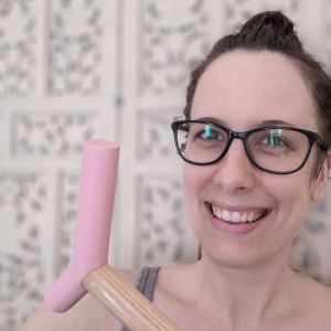 Brianne, a white woman in her mid-30s wearing black framed glasses and dark brown hair in a messy bun, smiles at the camera. She’s wearing a grey tank top, holding the pink epoxy handle of her blonde wood cane up beside her face and sitting in front of a blurred off-white decorative screen. The photo has been cropped close around Brianne’s face, shoulders and cane handle.