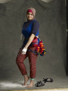 Anoushé, a woman wearing an athletic sports outfit, with a colorful leggings and a blue shirt. She is barefoot, has tape around her hand and fingers and around the elbow of her missing other arm. She wears a red and beige head scarf and is smiling slightly.