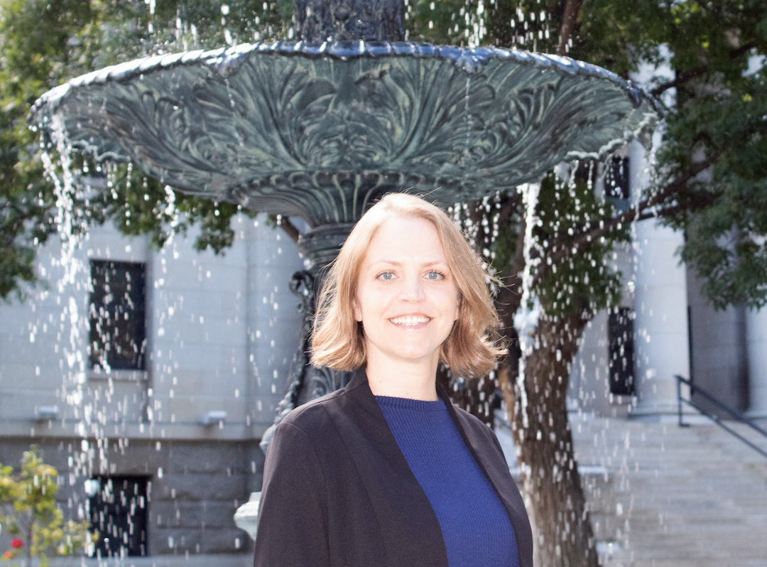 A woman with chin-long light brown hair stands in front of a fountain. She wears a blue shirt and a black blazer and smiles.