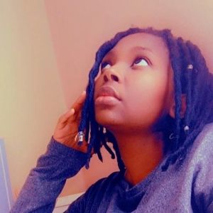 A black woman with chin-long black hair. She is looking to the side and has beautiful brown eyes and wears a grey sweater.