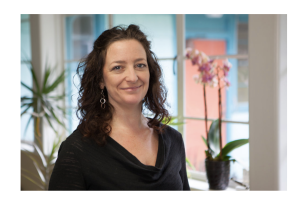 Kimby Maxson, a woman with curly brown hair is standing in a bright room with an orchid in the background.
