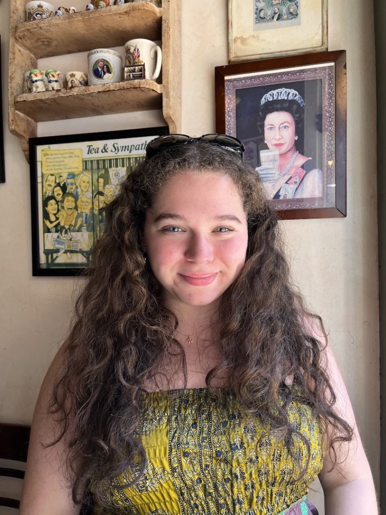 Lily, a young woman with long curly brown hair. She is wearing a yellow dress and has her sunglasses up on top of her head.
