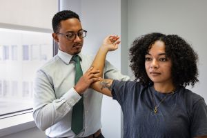 Jacques Courseault, a black man with black hair and black glasses. He wears a blue shirt and a green bow and lifts up the arm of a black woman with a tattoo on her arm in an examination room.