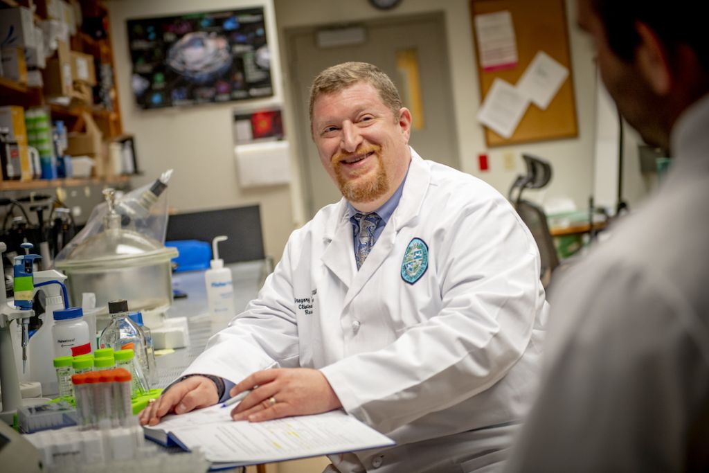 Gregory Bix, a white man with blonde hair and a red beard sits in a lab full of chemical flasks. He is wearing a white lab coat and is smiling towards a colleague.