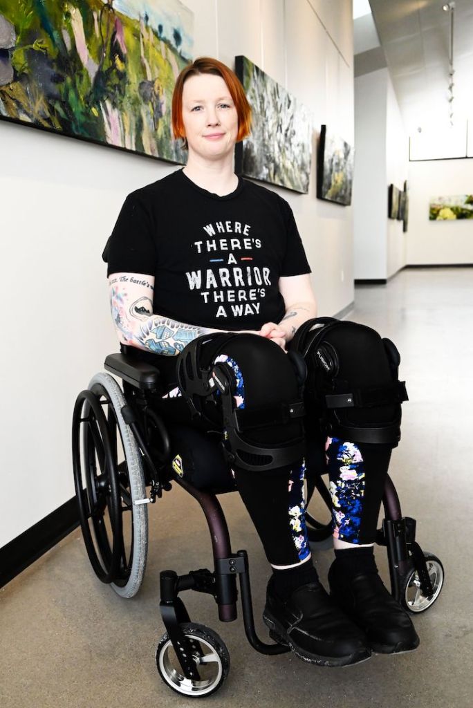 Pey Carter, a person with short red hair is in a gallery with beautiful art on the walls. They are in a wheelchair, wear braces and have many tattoos on their arms.
