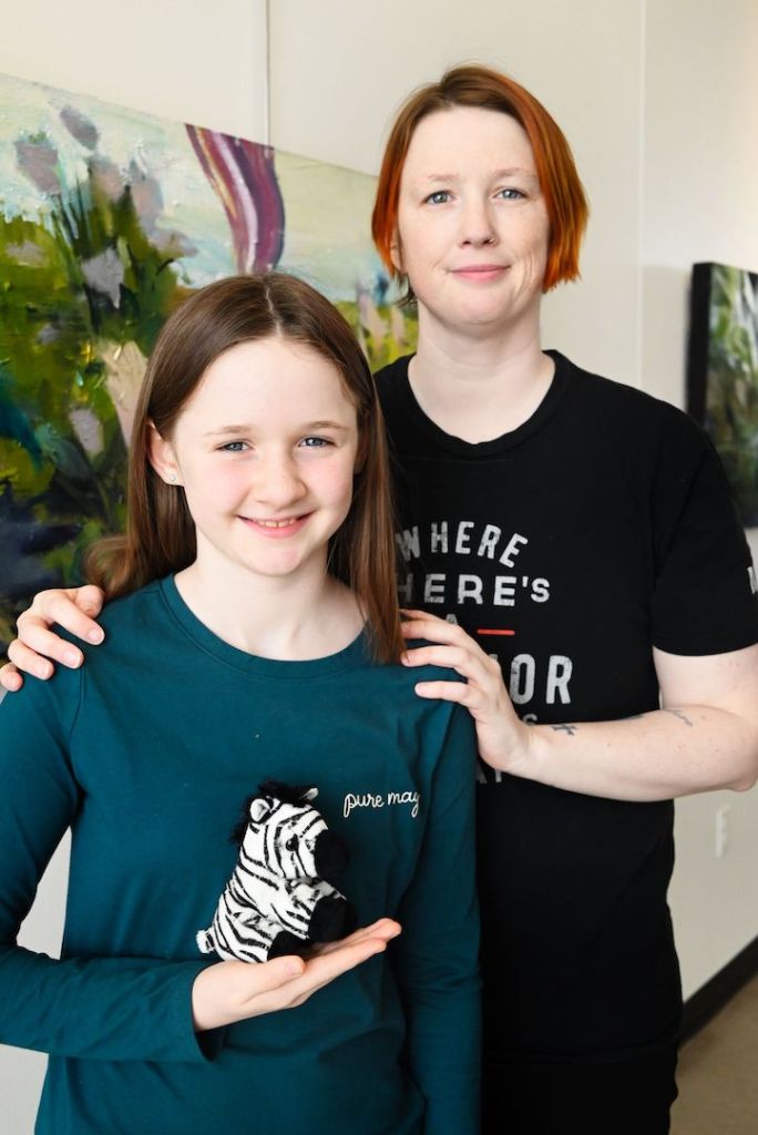 Pey and Abigail, mother and daughter stand next to one another. Abigail has long brown hair and is holding a zebra stuffed animal on her hand. Pey has short red hair and has their arms on Abigails shoulders