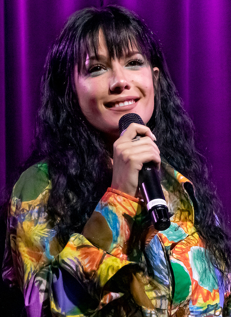 Halsey, a woman with curly brown long hair and a colorful outfit on a stage with a microphone in her hand.