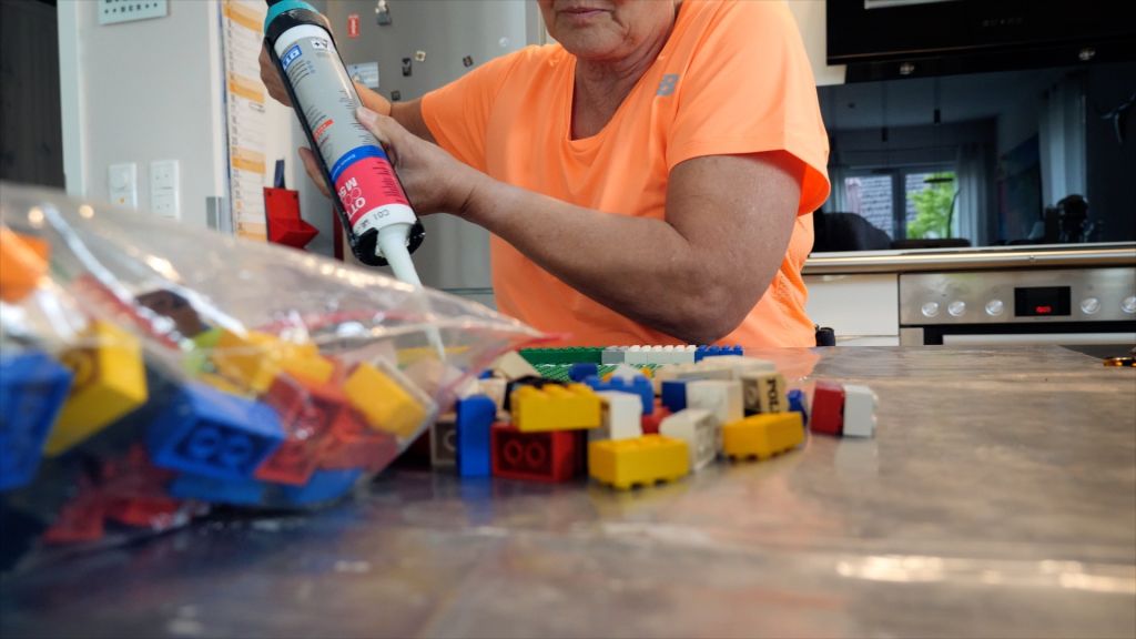 A person holding a big glue pistol sitting in a kitchen with a huge bag of colorful Lego in front.