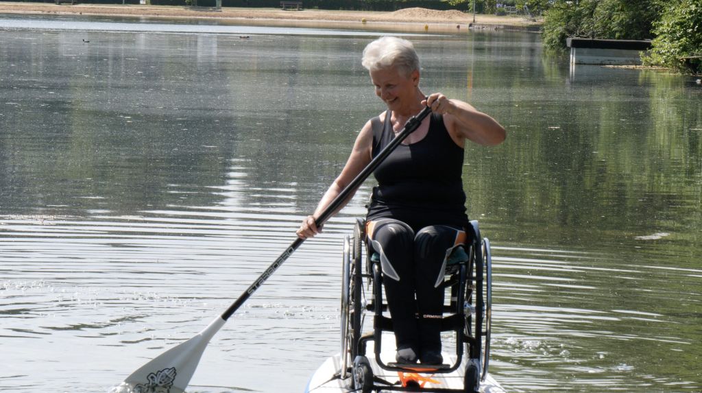 Rita, a woman with short grey hair sits in a wheelchair that's attached to a stand up paddle board on a lake. It's sunny and bright and she is smiling while holding a paddle.
