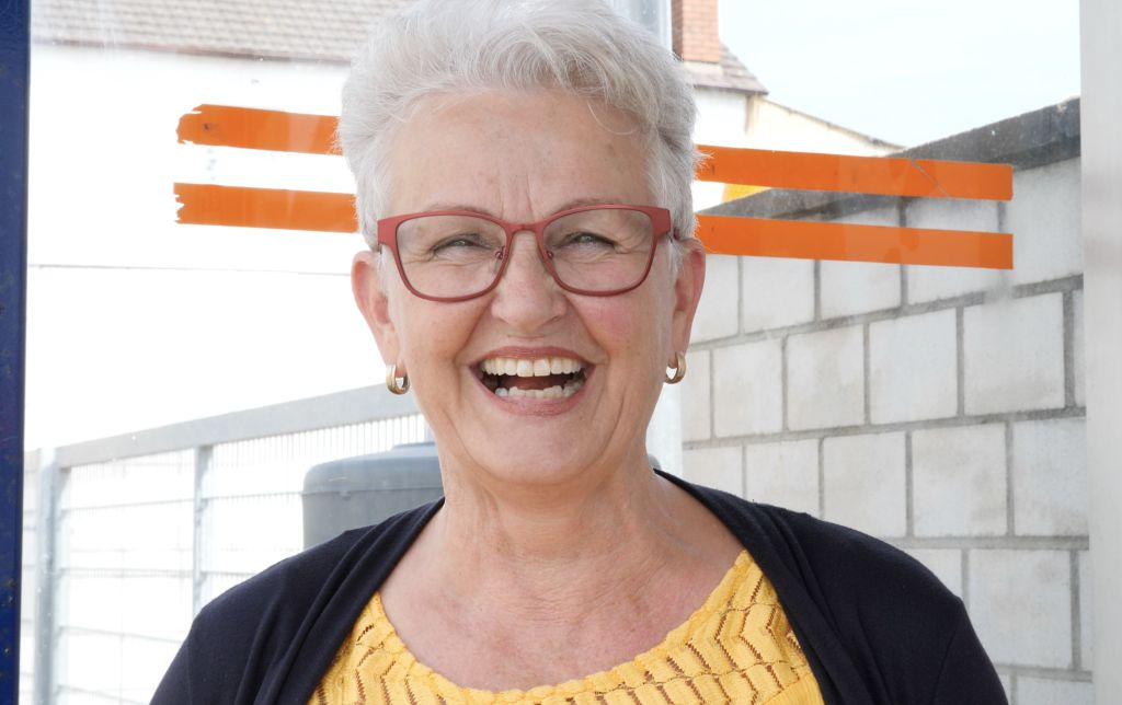 Rita, a woman with short grey hair and red glasses smiles brightly.