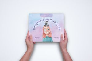 A pair of Caucasian hands holds the book Bendy Bones and Stretchy Skin: An Ehlers-Danlos Book. The cover of the book shows a watercolor painting of a young Caucasian girl wearing a teal shirt with a small stuffed animal zebra on her head. The background is a mix of soft pinks, purples, and blues.