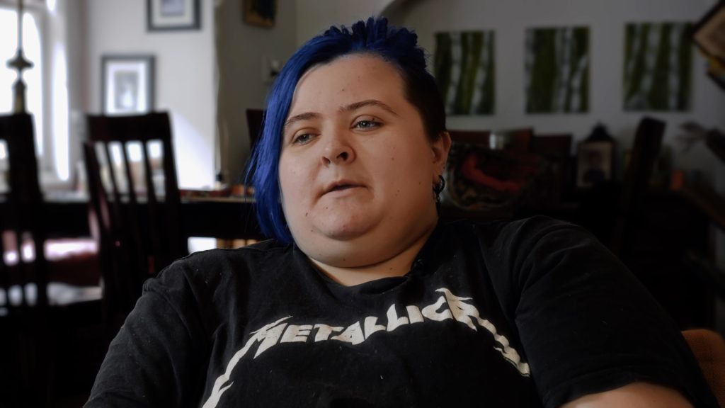 Jack, a young person with blue hair bound to a ponytail sits in a living room. They wear a Metallica shirt.
