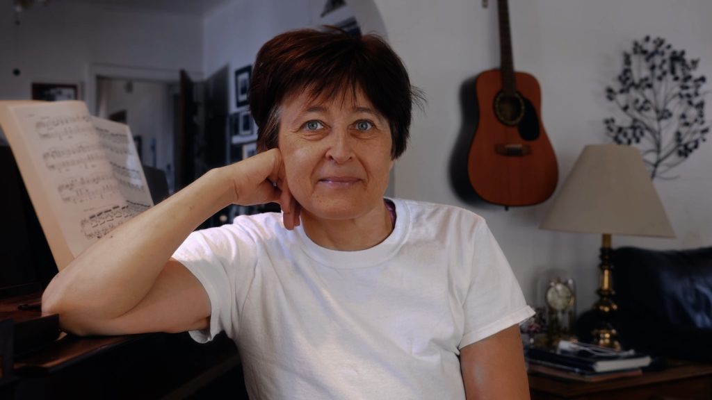 Maria, a person with short brown hair and blue eyes is sitting at a piano with their head propped on their hand.
