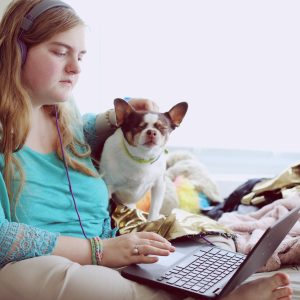 A woman with long blond hair in an aquamarine-colored top sits on a bed with a laptop between her legs. A small brown and white dog sits next to her.