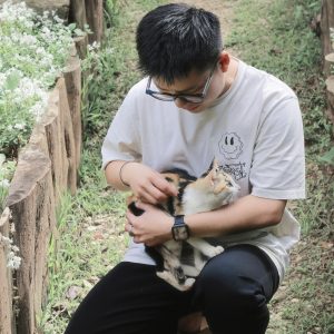 A man wearing a gray t-shirt, pants, and glasses crouches in the grass near a fence, holding a small calico cat.