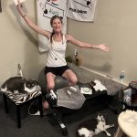 CHristie, a woman with long dark blonde hair bound to a pony tale sits in a corner of her room on a gymnastic ball with her arms spread out widely. She smiles. Next to her, on a zebra-colored chair, is a black and white cat napping. In front of her is a small desk with another black and white cat looking straight into the camera.