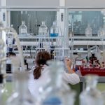 A view through some glass objects in a laboratory. In the background a person is pipetting things at a lab desk.