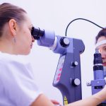 A patient and a doctor sitting on opposite sites of the table. The doctor examines the patient's eyes with a large machine.