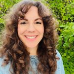 A white woman with curly brown hair in front of green bushes