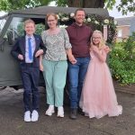 A family of four in front of a military vehicle. From left to right: A teen boy with blonde hair wearing a blue tie, a woman with redish hair and green pants, a man with a red shirt, and a young girl wearing a pink princess dress and a crown.