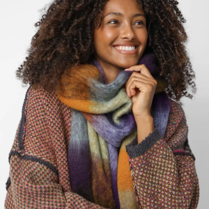 A women with dark coily hair wears a scarf that is multicolored (grass green, burnt orange, violet, brown, and more). The scarf is wide and long, allowing it to wrap around her neck at least once, with enough fabric for additional folds or wraps.