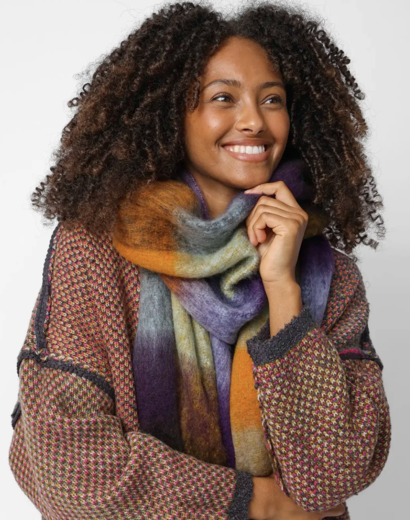 A women with dark coily hair wears a scarf that is multicolored (grass green, burnt orange, violet, brown, and more). The scarf is wide and long, allowing it to wrap around her neck at least once, with enough fabric for additional folds or wraps. 