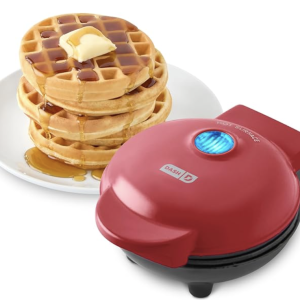 A small red waffle maker sits next to a stack of small waffles with butter and syrup on them.