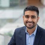 Jeevan, a brown man with black hair and a black beard is smiling brightly. He wears a blue suit.