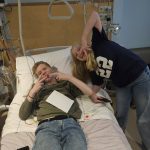 Two blonde children in a hospital. The boy is lying in a hospital bed while the girl is next to him making a victory sign with her hands.