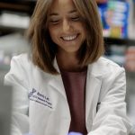 Amy Weintraub smiling while working in a laboratory, wearing a white Norris Lab research coat and blue gloves while handling materials at a lab bench surrounded by scientific equipment.