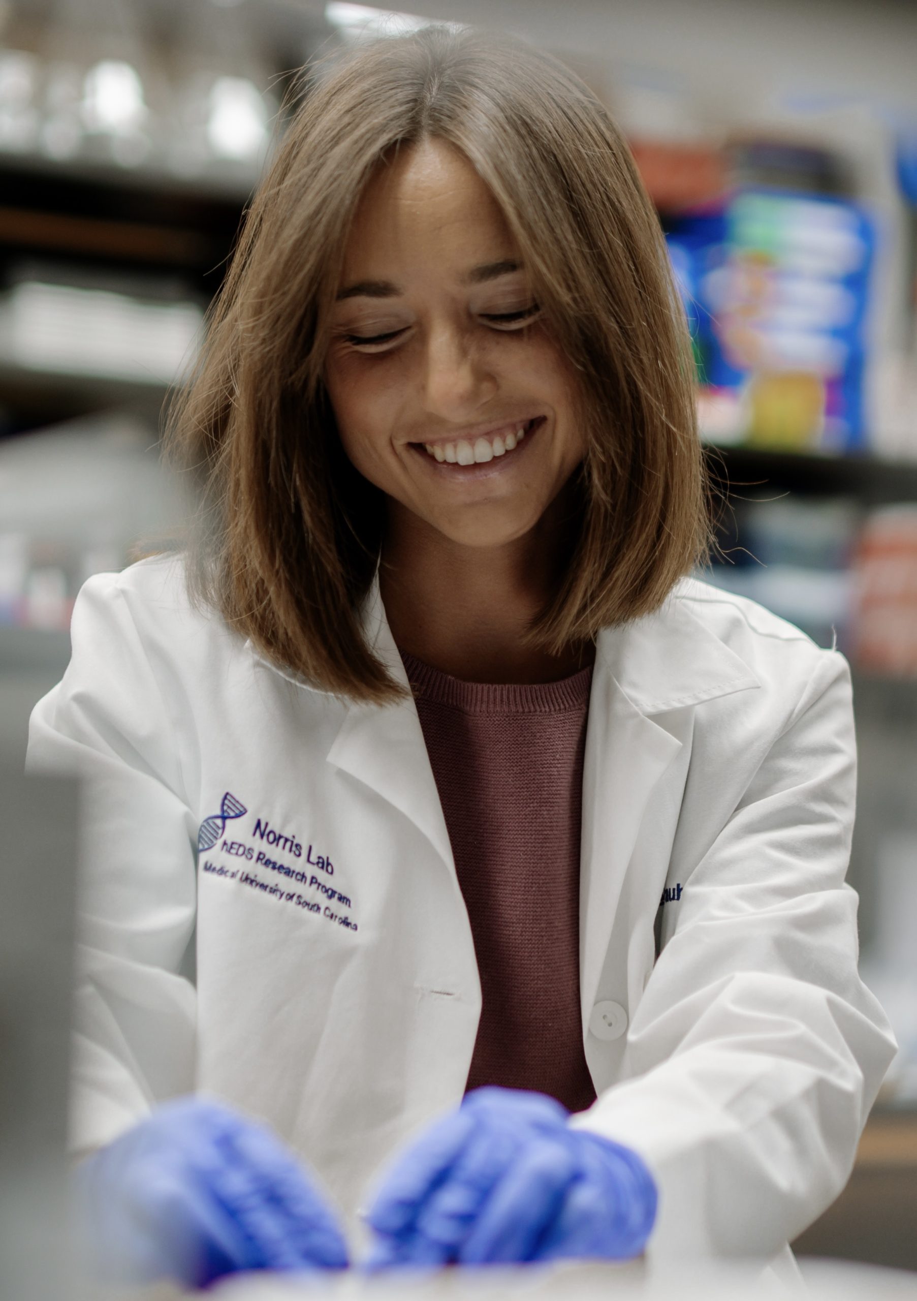 Amy Weintraub smiling while working in a laboratory, wearing a white Norris Lab research coat and blue gloves while handling materials at a lab bench surrounded by scientific equipment.