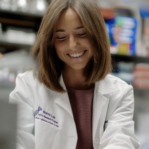 Amy Weintraub smiling while working in a laboratory, wearing a white Norris Lab research coat and blue gloves while handling materials at a lab bench surrounded by scientific equipment.