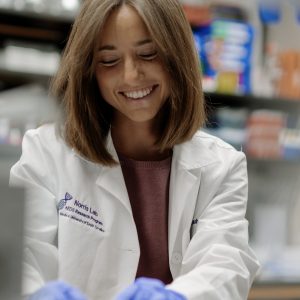 Amy Weintraub smiling while working in a laboratory, wearing a white Norris Lab research coat and blue gloves while handling materials at a lab bench surrounded by scientific equipment.