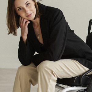 Tayler Goectau seated in a wheelchair, wearing a black blazer and light-colored pants, resting her head on her hand and smiling softly against a minimalist indoor background.