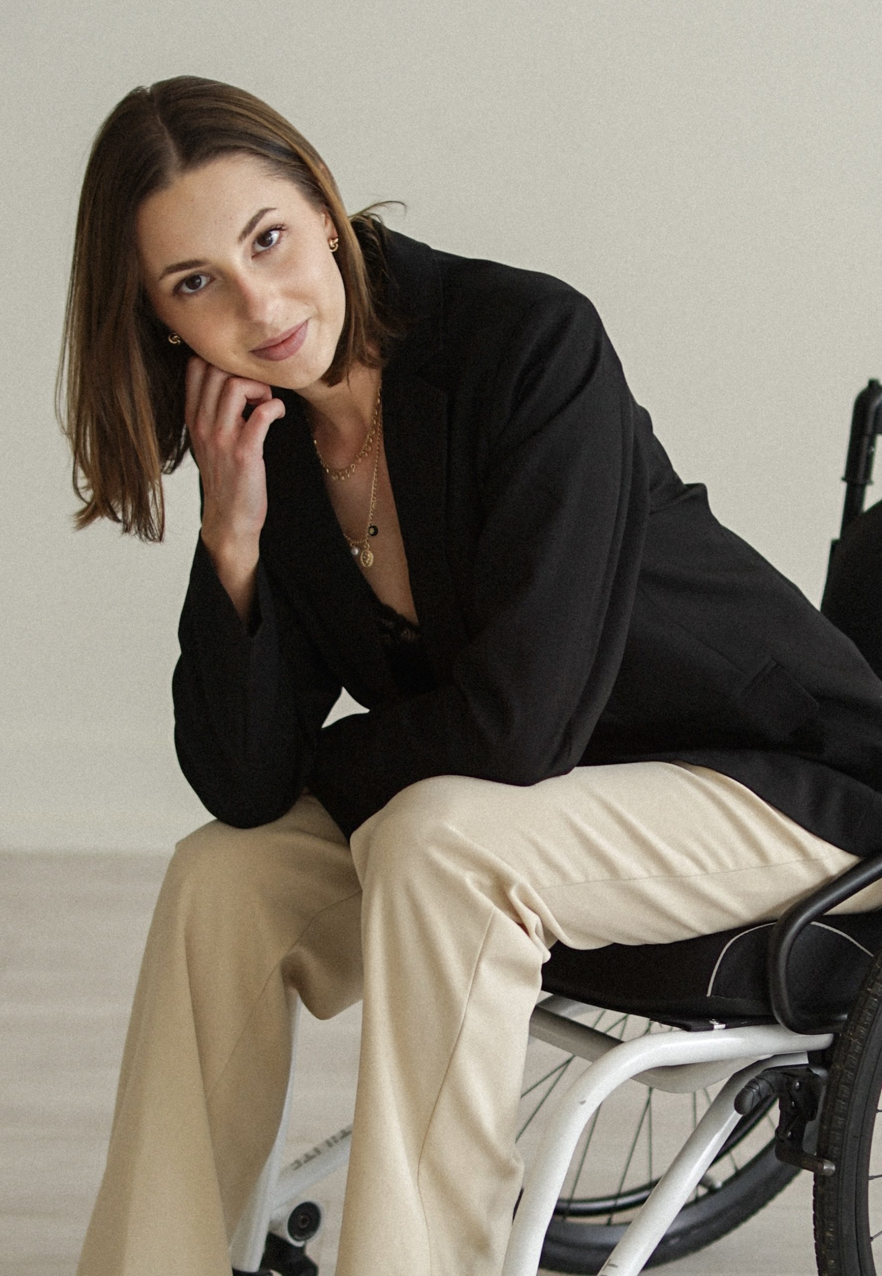 Tayler Goectau seated in a wheelchair, wearing a black blazer and light-colored pants, resting her head on her hand and smiling softly against a minimalist indoor background.