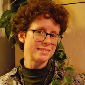 Portrait of Jan with short, curly brown hair wearing glasses and a dark turtleneck layered under a patterned shirt, smiling slightly. They are indoors, with a leafy green plant behind them and warm-toned lighting that gives the scene a cozy feel.
