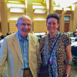 Dr. Rodney Grahame and Jan Groh smiling together at the EDNF conference in Rhode Island in 2013, standing in a warmly lit conference hall. Dr. Grahame is wearing a light beige blazer over a blue striped shirt and brown trousers, while Jan Groh is wearing a patterned dress with a long scarf, glasses, and a wrist brace, holding a blue water bottle.
