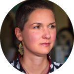 Portrait of Karina Sturm with short hair looking slightly to the side, wearing a patterned top and a gold leaf-shaped earring, with a softly blurred indoor background.