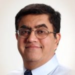 Portrait of Dr. Chopra, a middle-aged man with short dark hair and glasses, wearing a light blue shirt and tie, facing the camera with a slight smile against a neutral background.