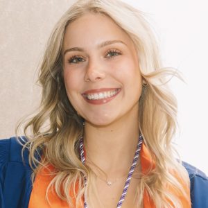 Professional headshot of Hailee Munno smiling at the camera with shoulder-length blonde wavy hair, wearing a blue graduation gown with an orange stole and honor cords against a soft neutral background.