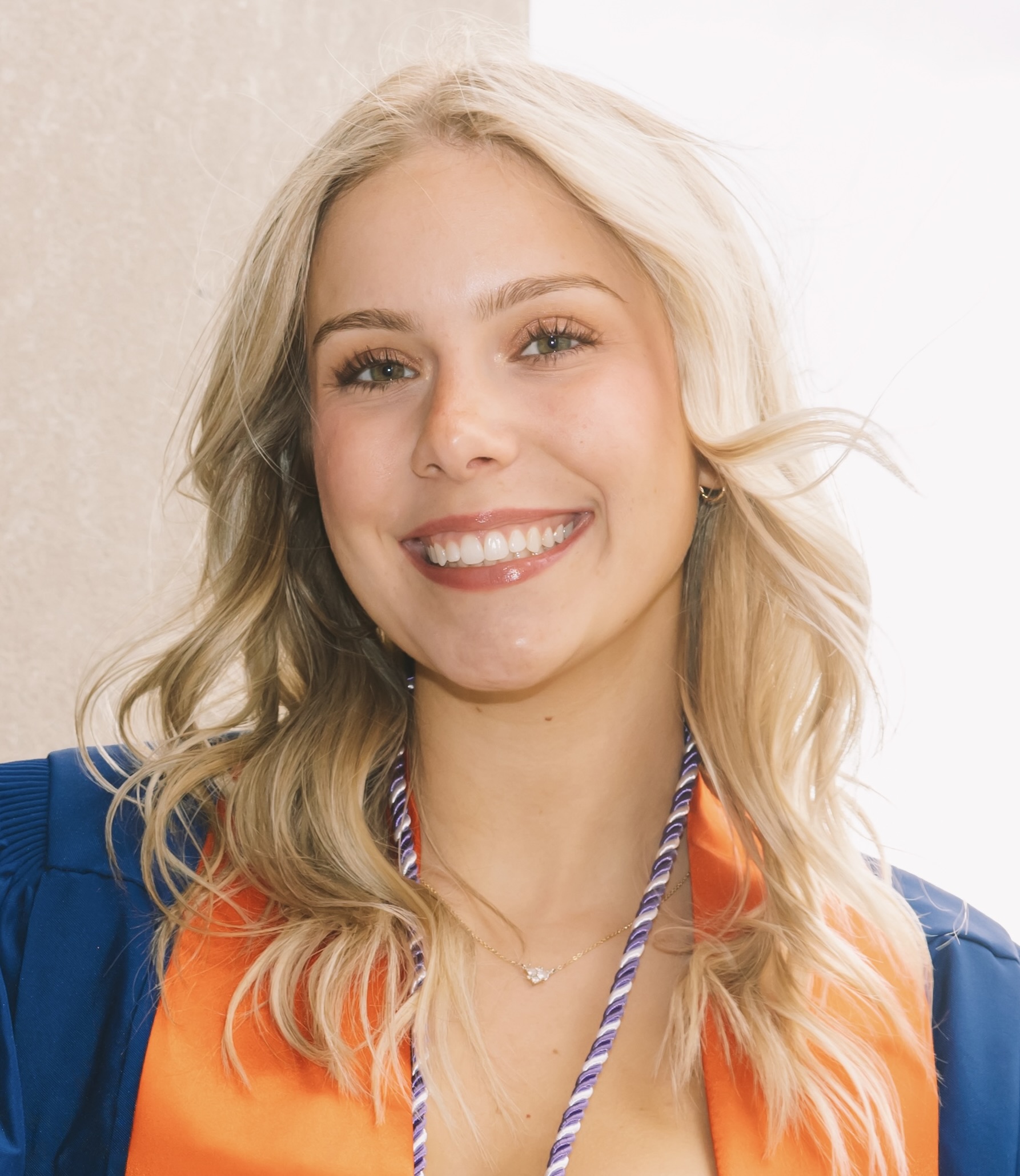 Professional headshot of Hailee Munno smiling at the camera with shoulder-length blonde wavy hair, wearing a blue graduation gown with an orange stole and honor cords against a soft neutral background.