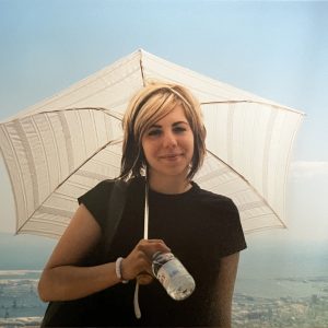 Jacqueline, in a black t-shirt, with blond and brown highlighted hair, standing holding a sun umbrella and a bottle of water, backlight by sunlight. The background seems as though she is high up on a mountain with a Japanese city below.