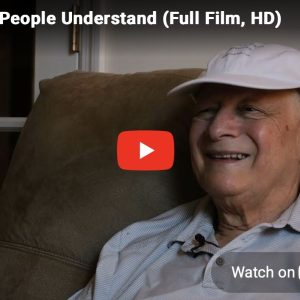 YouTube video thumbnail for the documentary Where People Understand – The New Short Doc Highlighting John Ferman’s Legacy. John Ferman is seated in a comfortable armchair, smiling during an interview. He is wearing a white baseball cap and a light-colored polo shirt with a lavalier microphone clipped near the collar. The thumbnail includes the documentary title at the top, a large red YouTube play button in the center, and a "Watch on YouTube" button in the lower-right corner. A glass-paneled door and indoor home setting are visible in the background.