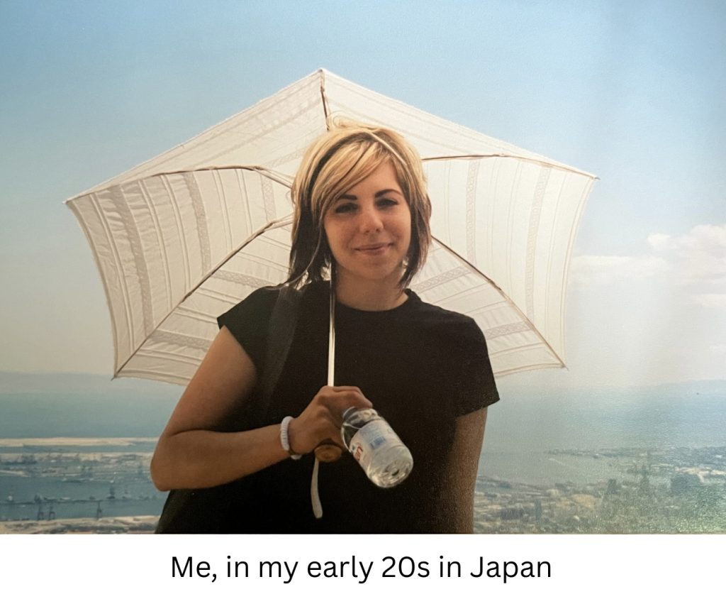 Jacqueline in her early 20s in Japan. She has a black t-shirt on, blond and dark brown highlighted hair and is holding a cream sun umbrella being backlit by the sun. She is also holding a water bottle. She is high up on a mountain with a Japanese city visible far below. She has a soft smile.