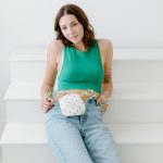 Tayler sits on white stairs wearing a green sleeveless top and ripped jeans, with an ostomy pouch visible at her waist. Small white flowers are tucked into the waistband of her jeans around the ostomy pouch. She looks toward the camera with a calm, confident expression, portraying everyday life with an ostomy.