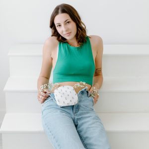 Tayler sits on white stairs wearing a green sleeveless top and ripped jeans, with an ostomy pouch visible at her waist. Small white flowers are tucked into the waistband of her jeans around the ostomy pouch. She looks toward the camera with a calm, confident expression, portraying everyday life with an ostomy.