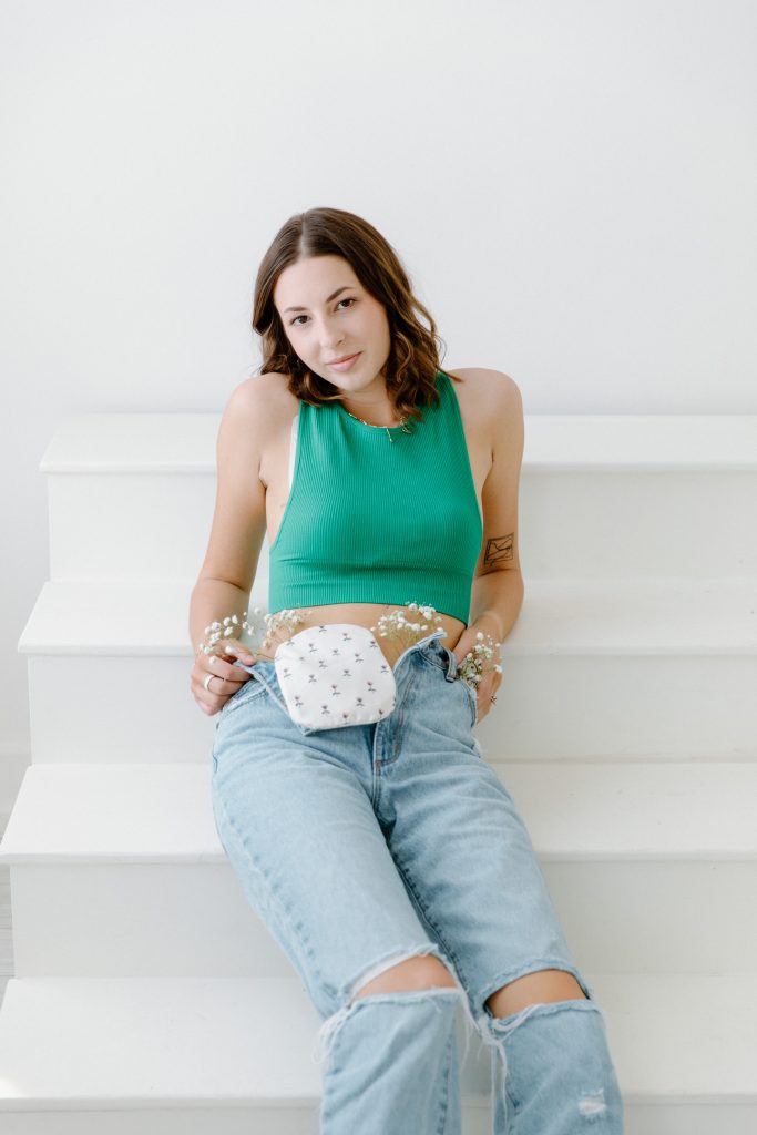 Tayler sits on white stairs wearing a green sleeveless top and ripped jeans, with an ostomy pouch visible at her waist. Small white flowers are tucked into the waistband of her jeans around the ostomy pouch. She looks toward the camera with a calm, confident expression, portraying everyday life with an ostomy.