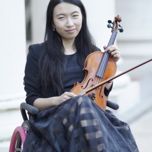 Amy Wang-Hiller seated in a wheelchair, holding a violin and bow, wearing a black dress and smiling at the camera.